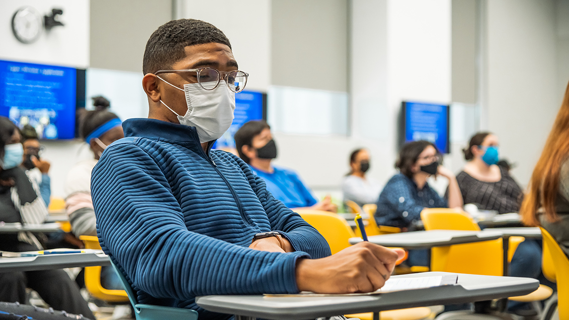 A student wearing a face covering taking notes during a class.