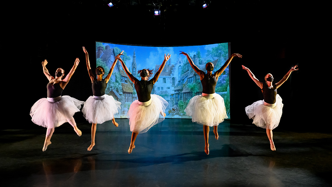 5 dancers in black shirts and tutus leap into the air onstage, with their arms stretched above their heads.
