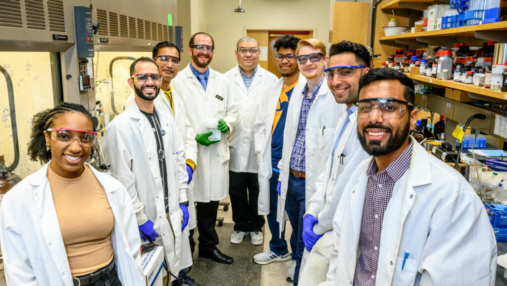 Dr. Mitch Croatt and 9 students standing in a narrow lab aisle.