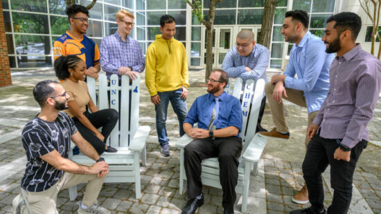 Dr. Croatt and his lab members stand around two white rocking chairs outside of the Sullivan Science building.
