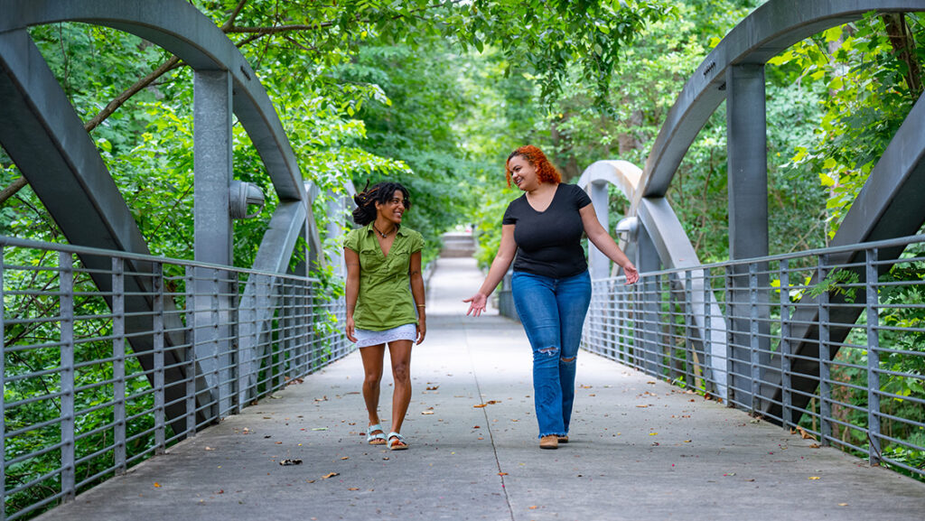 Two college students walk across a footbridge surrounded by trees.