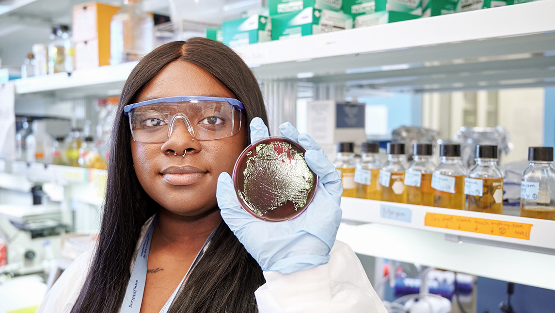 Student in a lab holds up a petri dish to the camera