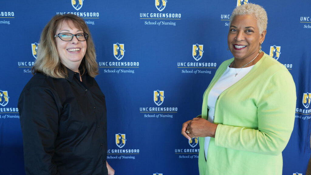 Two faculty members pose in front of UNCG logo wall