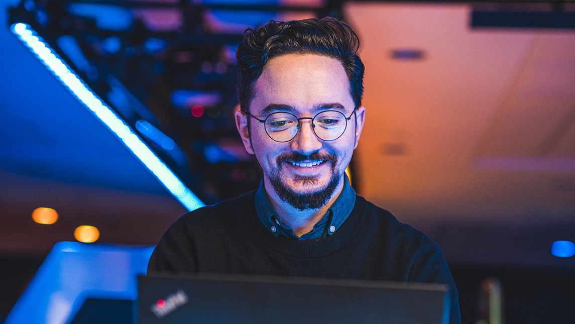 Bespectacled man smiles from behind his laptop