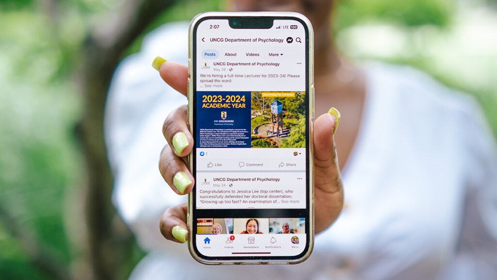 Someone holding out a smartphone open to UNCG's Department of Psychology's Facebook page