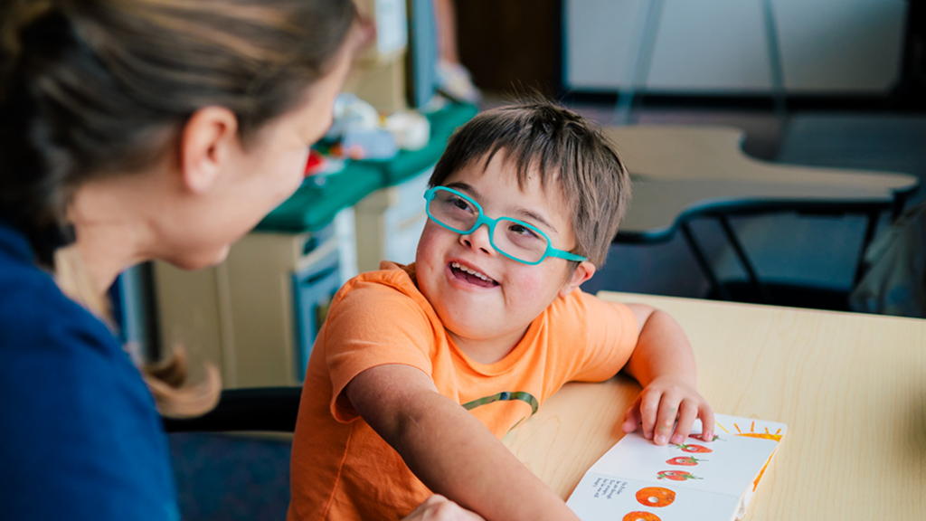 Teacher facing young child wearing blue glasses and reading book together