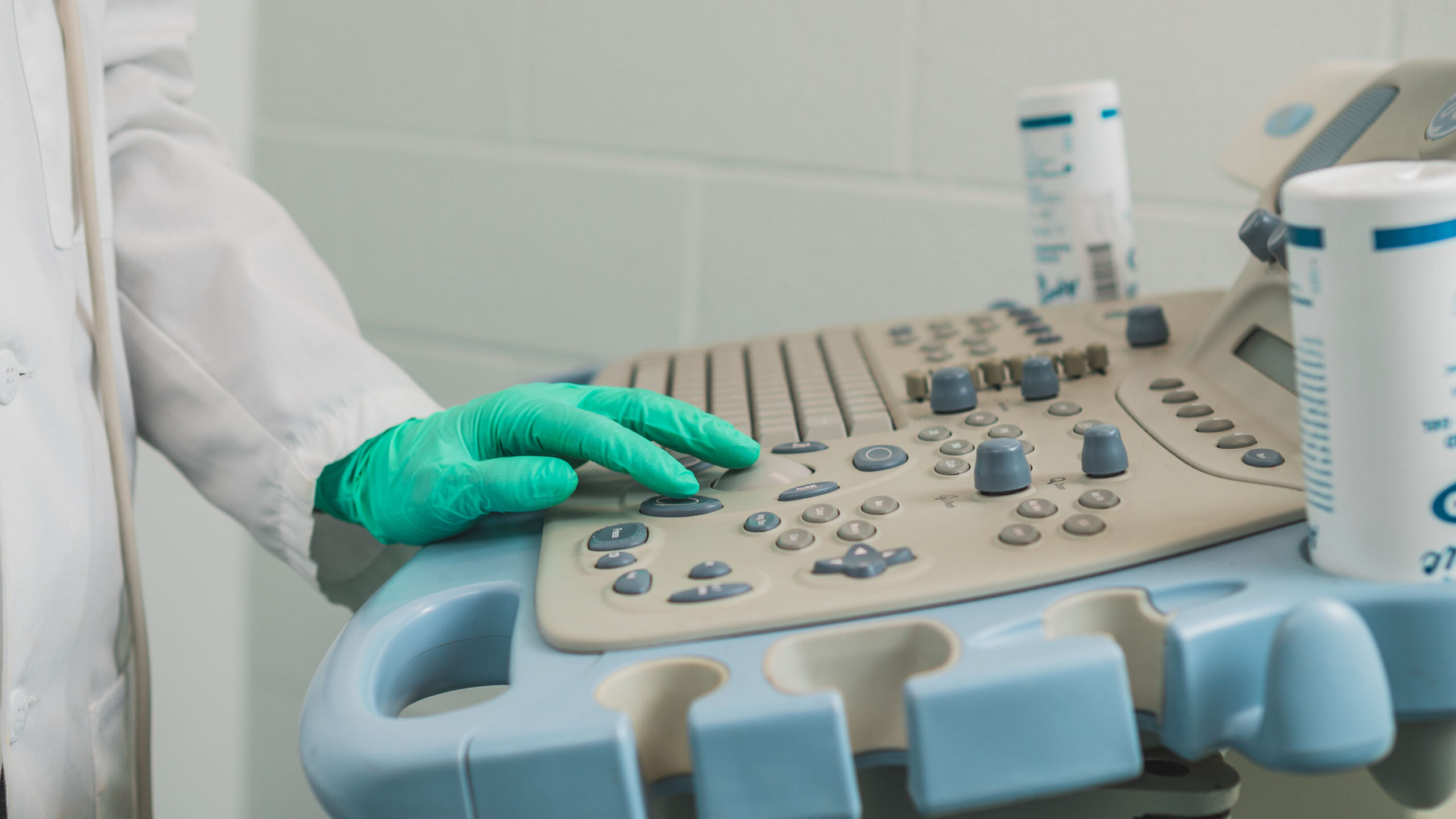 Person in lab coat and green gloves has hands on keyboard of echocardiogram machine