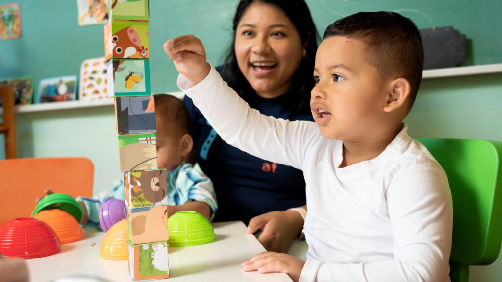 Latine toddler stacks blocks with expression of excitement while early childhood education professional cheers him on in a classroom setting.