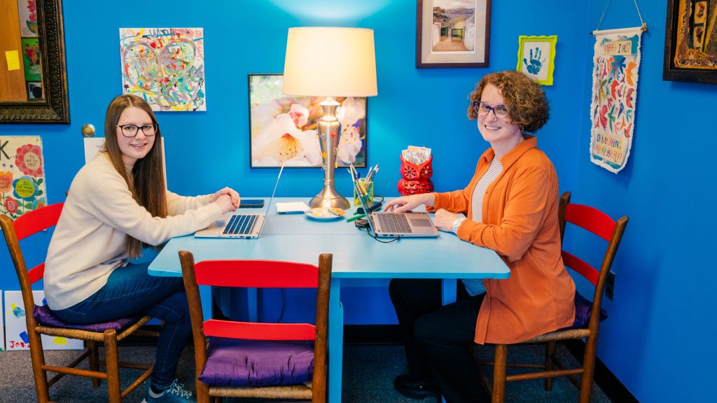Undergraduate Kaya McDowell and Kim Cuny pose at a table with their laptops