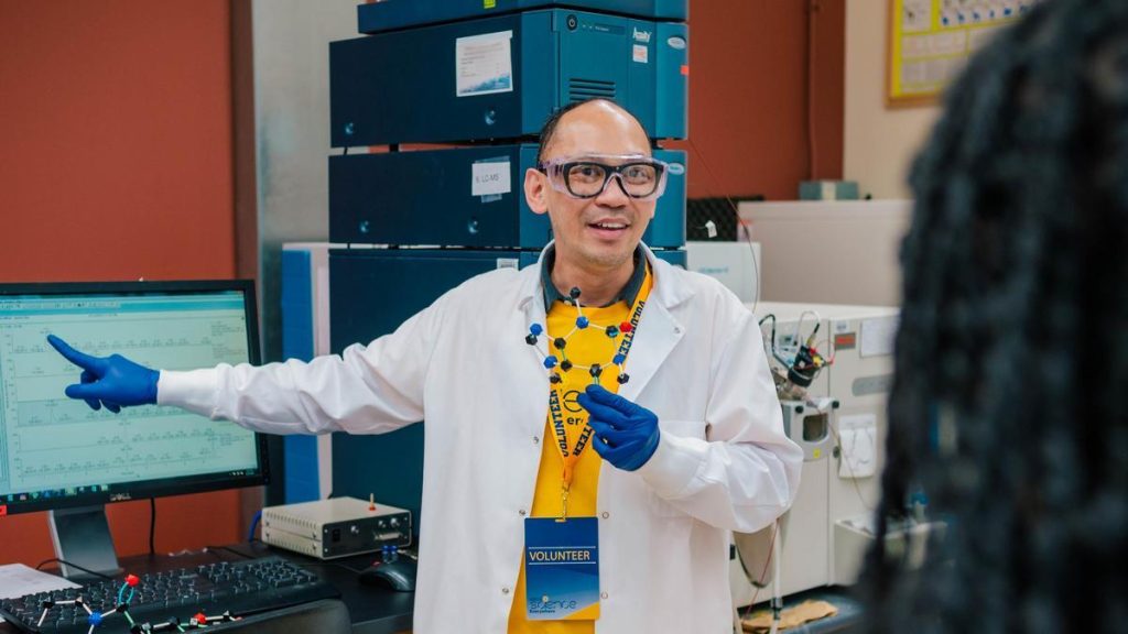 A man wearing a volunteer lanyard points at a screen as he wears a lab coat and gloves