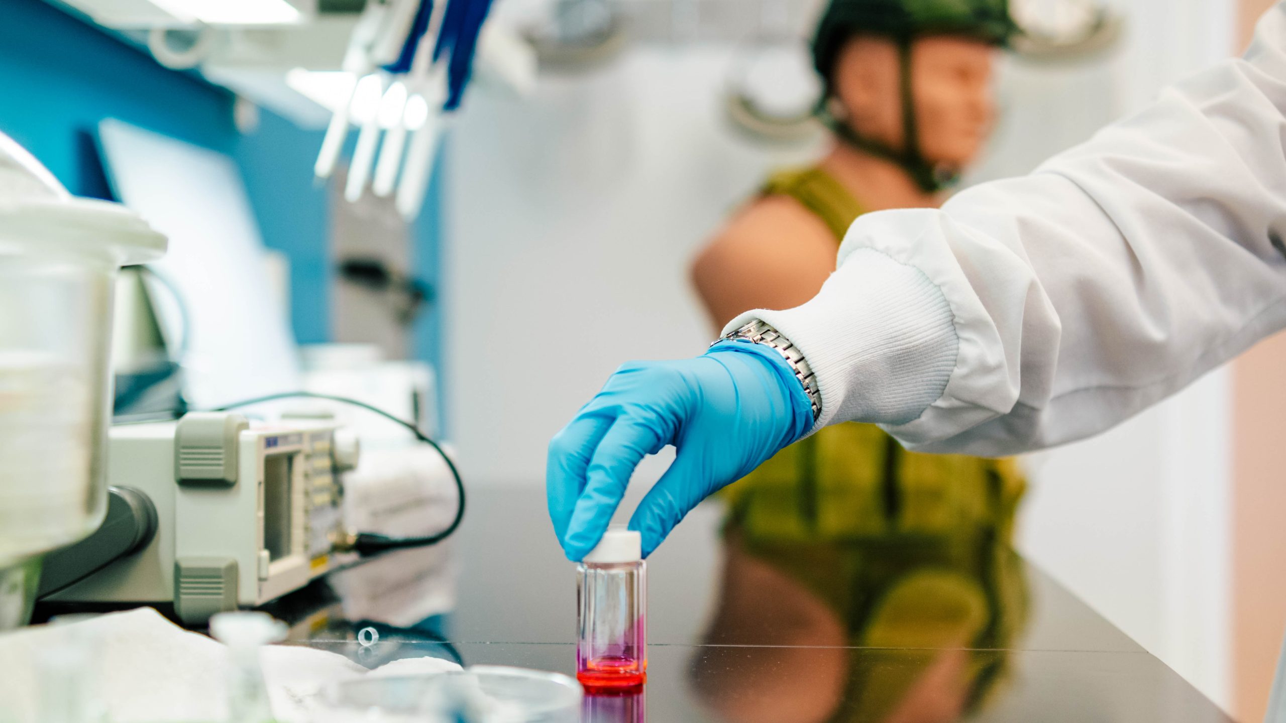 A gloved hand places a vial of a glowing red substance on a table