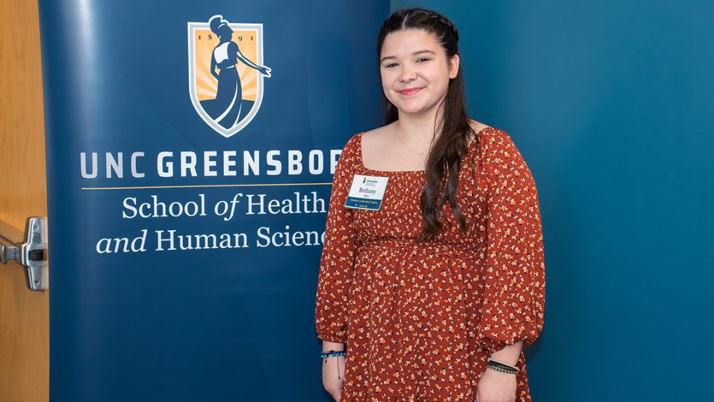 Bethany Allen standing in front of a blue backdrop featuring the UNC Greensboro School of Health and Human Sciences logo and text.