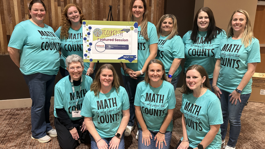 Group of people wearing matching turquoise T-shirts that read ‘Math Counts’ posing together indoors. They are standing and kneeling around a sign on an easel that says ‘Featured Session’ with logos for NCTM and other organizations. The background includes wooden panels and a carpeted floor.