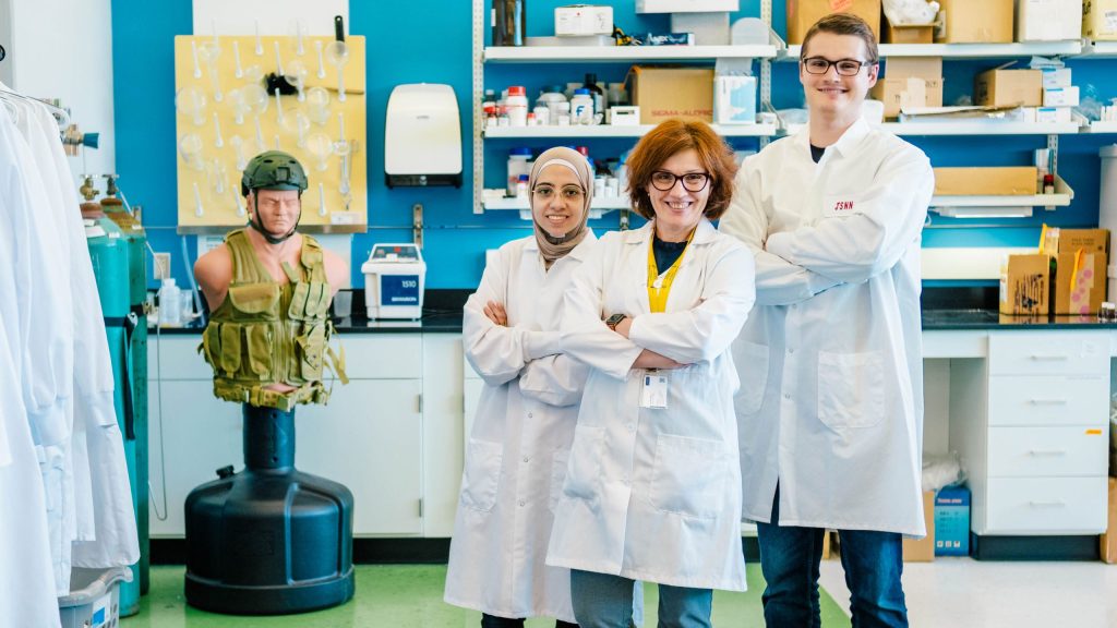 Three individuals wearing white lab coats stand in a laboratory with arms crossed. Behind them are shelves with scientific equipment, boxes, and supplies. To the left, there is a mannequin torso dressed in a military-style vest and helmet, positioned on a black stand. The background includes a blue wall, a mounted paper towel dispenser, and various lab instruments.