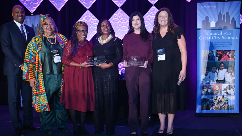 Guilford County School members standing on a stage holding clear award plaques. They are dressed in formal and colorful attire. Behind them is a backdrop with geometric lighted panels, and to the right is a vertical banner that reads ‘Council of the Great City Schools’ with images of children and a city skyline graphic.