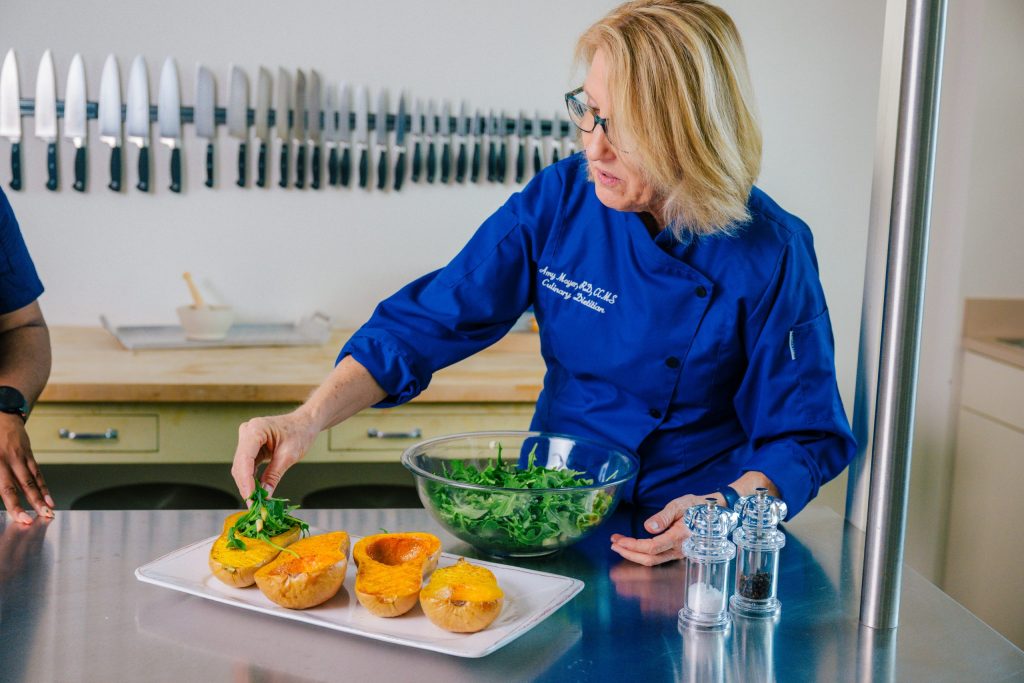 Person in a blue chef coat preparing food on a stainless steel counter, adding greens to halved roasted squash with a bowl of leafy greens nearby. A row of kitchen knives is mounted on the wall in the background.