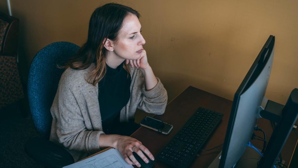 Person working at a desk on a desktop computer, with a keyboard, mouse, phone, and documents nearby.