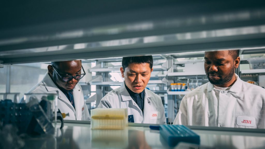 Researchers wearing white lab coats work together in a laboratory setting, surrounded by scientific equipment and supplies, including pipette tip boxes and containers on a bench.