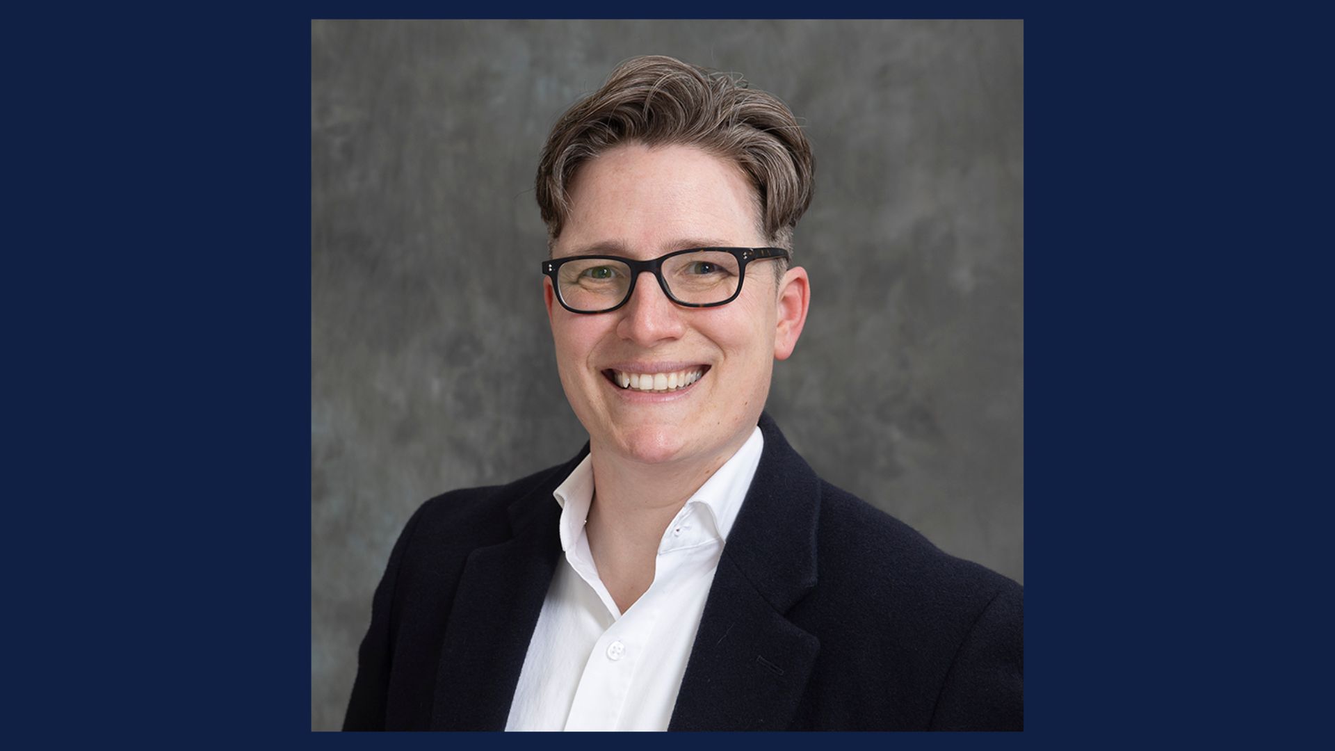 Dr. Jennifer Feather headshot wearing a dark blazer and white shirt against a gray studio backdrop.