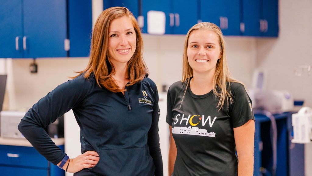 Two people standing in a laboratory workspace with blue cabinets and scientific equipment in the background.