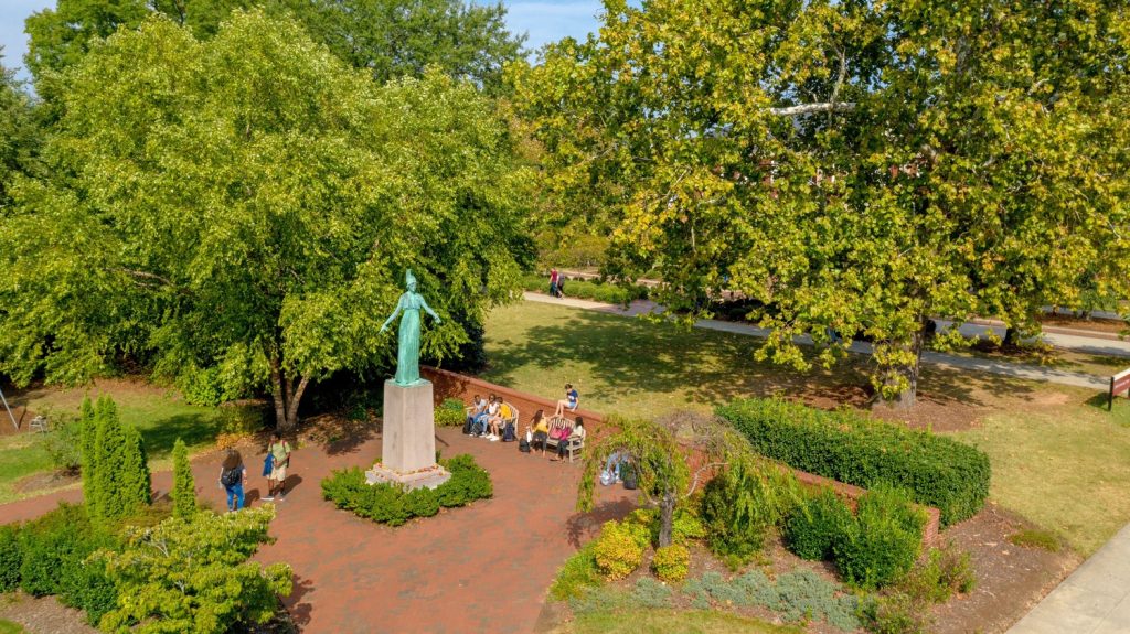 An aerial view of campus featuring the minerva statue at the center, surrounded by brick pathways, trees, and landscaped greenery, with a small group of people gathered near the base.