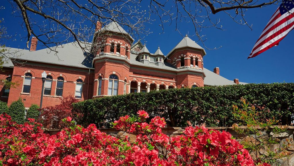 The College of Arts & Sciences building with towers and arched windows, framed by blooming pink flowers and an American flag against a bright blue sky.