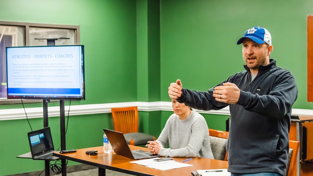 Jeff Milroy standing and gesturing during a presentation while another person sits at a table with a laptop; a screen displays slides in a green-walled classroom.