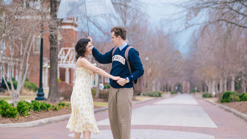 Two people holding hands and walking on a brick campus walkway, with one holding a clear umbrella overhead and historic campus buildings and trees lining the path.