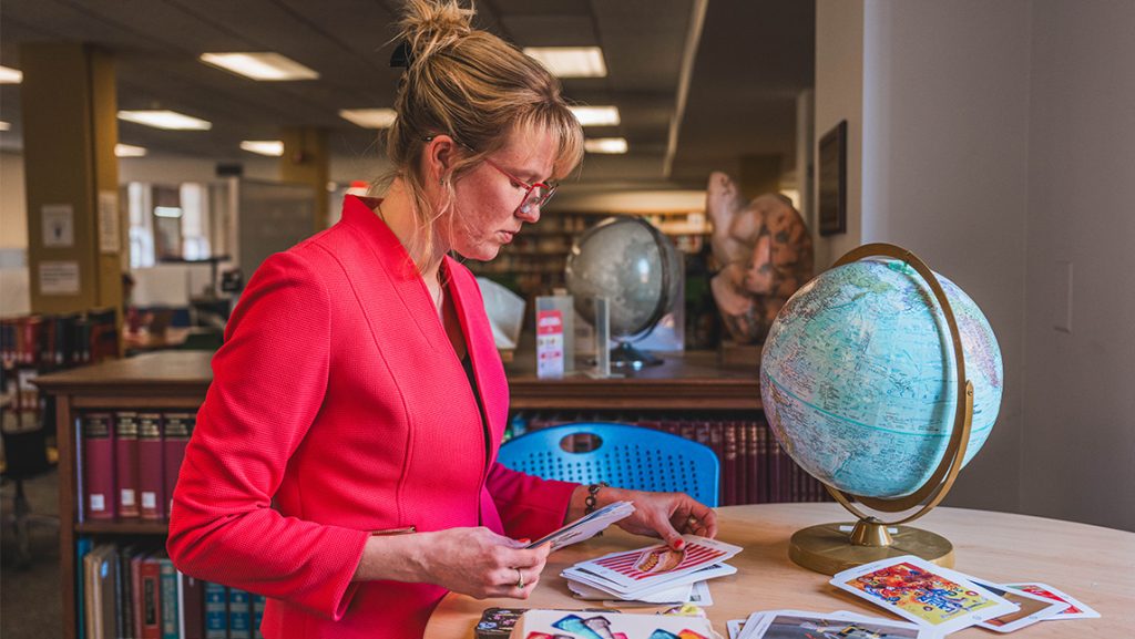 A person in a bright pink jacket sorting through printed images on a table in a library, with a globe and bookshelves visible in the background.