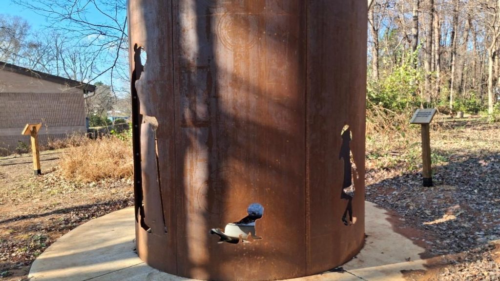 A large cylindrical outdoor sculpture with human silhouettes cut into its metal surface, installed along a wooded pathway with informational plaques nearby.