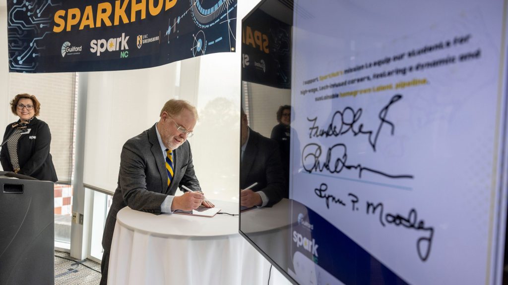Person signing a document at a tall table during a SPARKhub event, with a large display showing signatures and branding materials visible in the background.