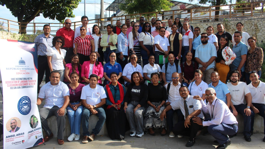 Large group photo taken outdoors on a paved area, showing dozens of participants arranged in rows in front of a stone wall and railing. A mix of individuals are standing and sitting, many wearing name tags, casual or business‑casual clothing. At the left side of the image, a tall event banner displays logos, text, and photos related to a professional workshop or community program.