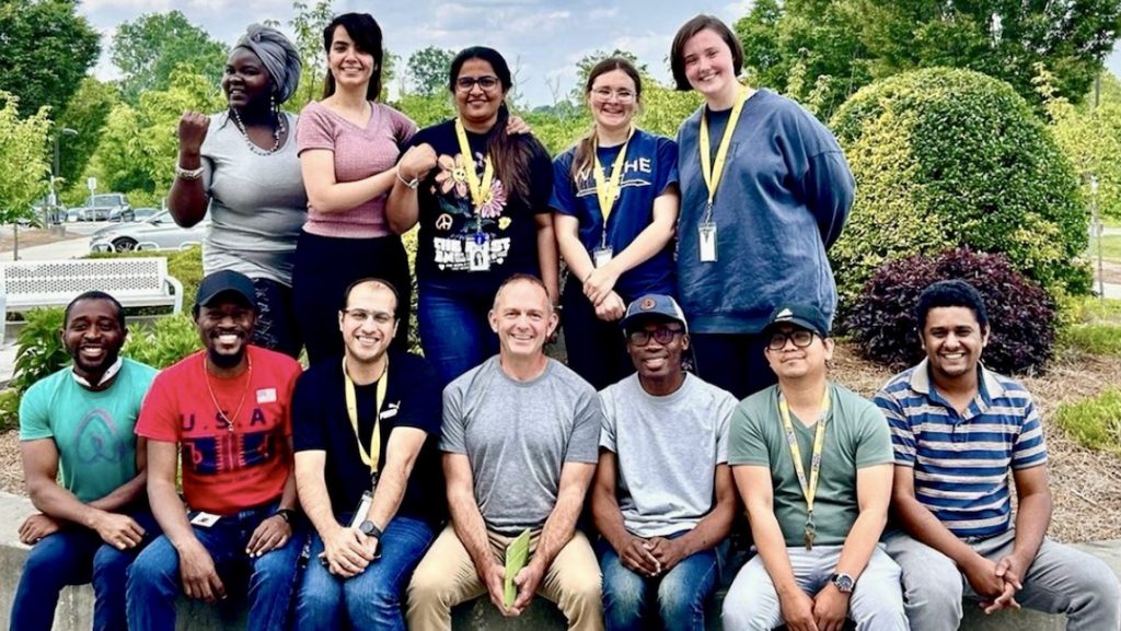 Group of eleven people posing outdoors in front of landscaping with trimmed bushes and trees. Five individuals stand in the back row wearing lanyards, and six individuals sit or kneel in the front row, all dressed casually and smiling during a group event or program gathering.