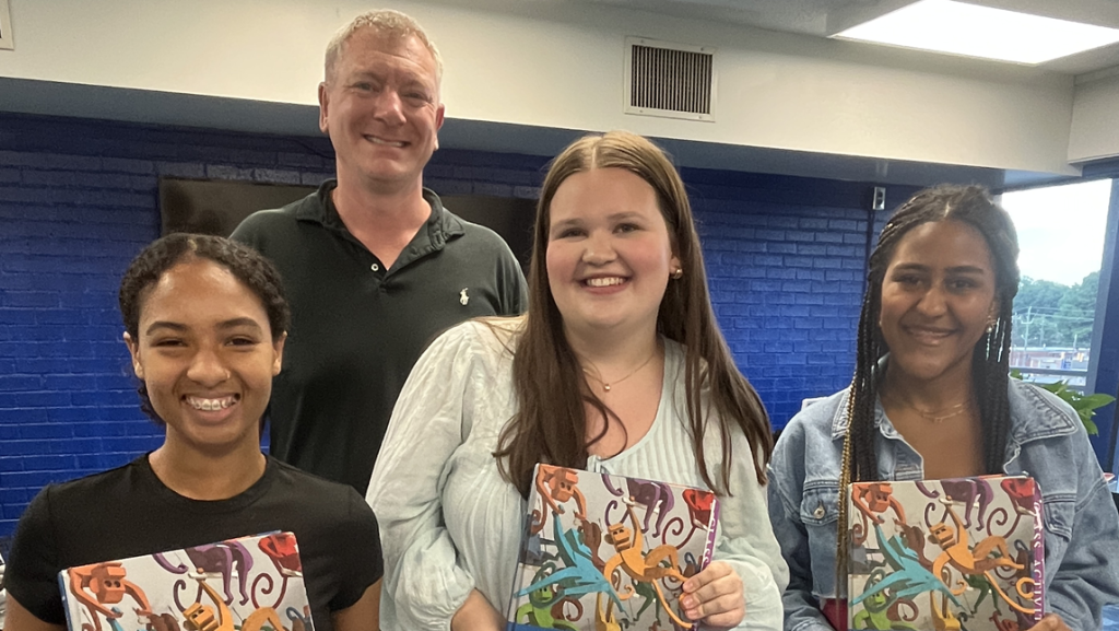 Four people standing indoors in front of a blue wall, each holding a colorful CALO book, with another individual standing behind them.