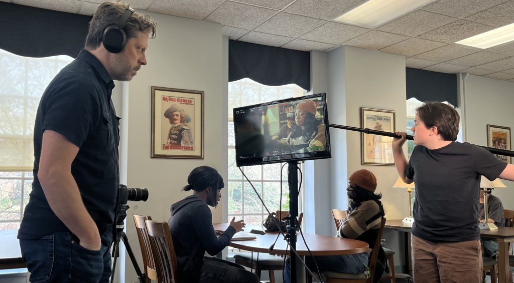 Film production students working in a classroom, with one person operating a camera, another holding a boom microphone, and a monitor displaying the scene being recorded while others sit at tables in the background.