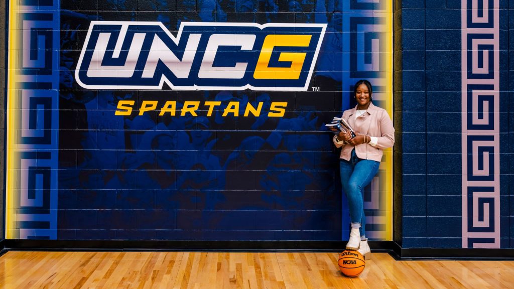 Lexi Kier standing in a gymnasium hallway holding notebooks and papers, leaning against a wall featuring the large UNCG Spartans logo, with a basketball placed under one foot.
