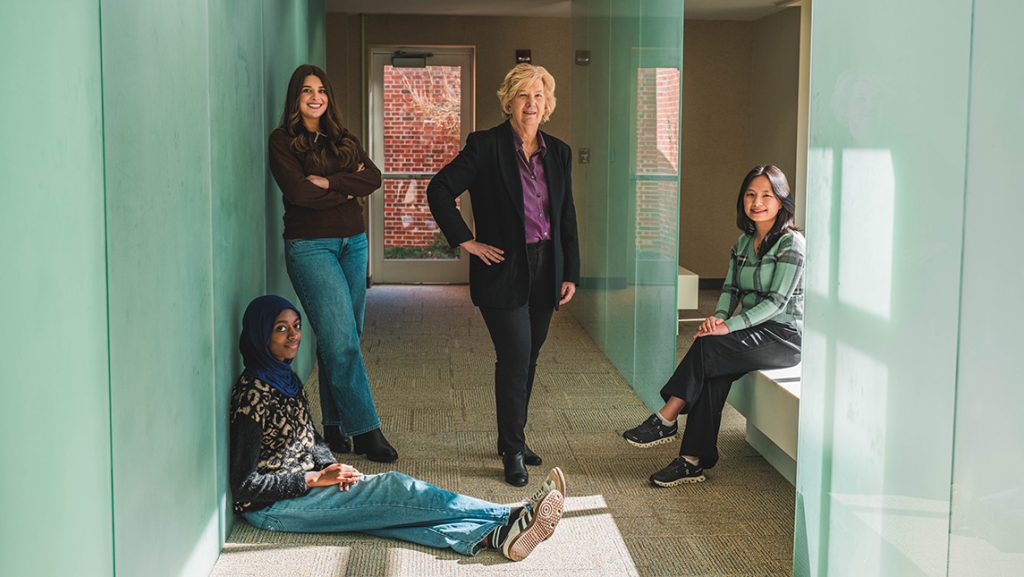 Group of four people in a bright hallway with green-tinted glass walls, positioned casually— Susan Keane is standing with a student, while one is seated on a bench, and one sitting on the floor—engaged in conversation in a modern academic building.